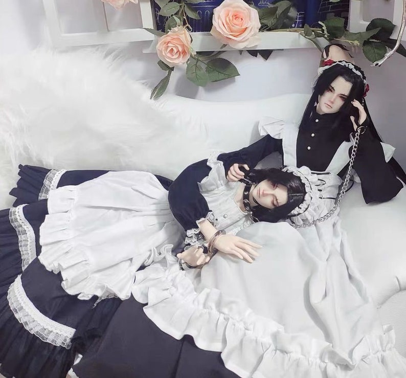 May include: Two dolls wearing maid outfits with black and white ruffles. One doll is wearing a black choker with a silver chain. The dolls are lying on a white bed with a white fluffy blanket.