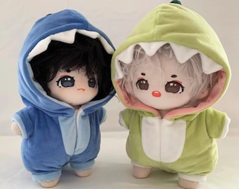 May include: Two plush toys, one blue and one green, are dressed in hooded outfits. The blue toy has a shark-like mouth on the hood and the green toy has a dinosaur-like mouth on the hood. Both toys have painted faces and black hair.