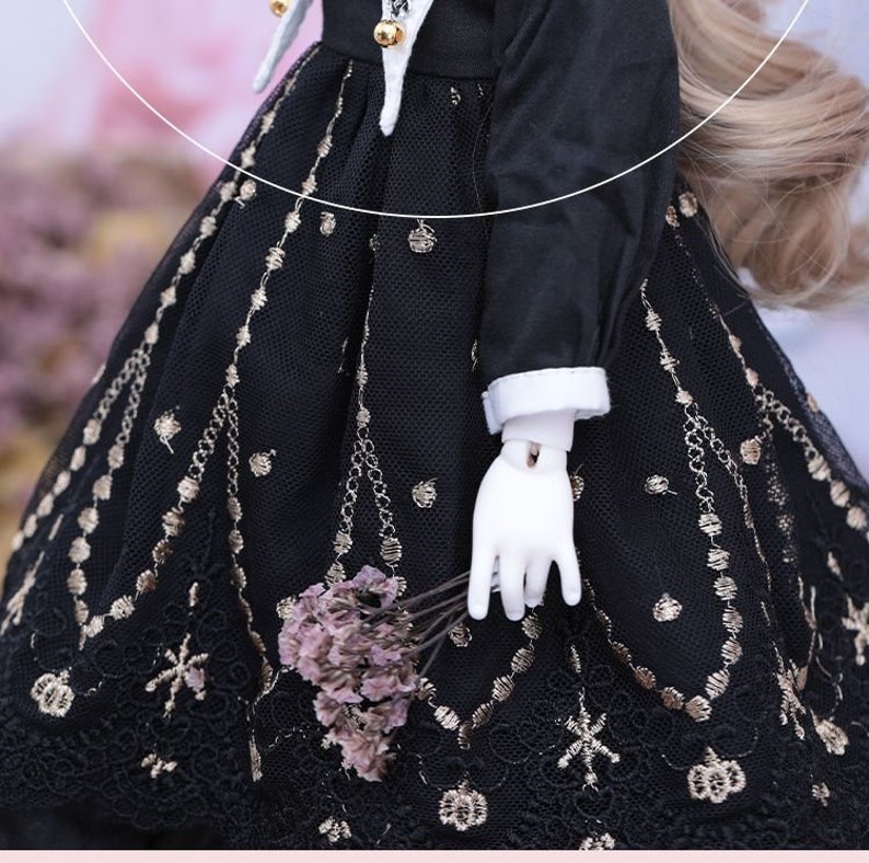 May include: A black doll dress with gold embroidery and lace detailing. The dress features a white collar and cuffs, and the doll is holding a small bouquet of dried flowers. The dress has a vintage aesthetic.