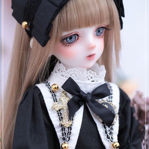 May include: A detailed portrait of a doll with long, light brown hair and blue eyes. The doll wears a black dress with a white collar and a black bow. A gold cross pendant and a black headband with gold accents complete the look.