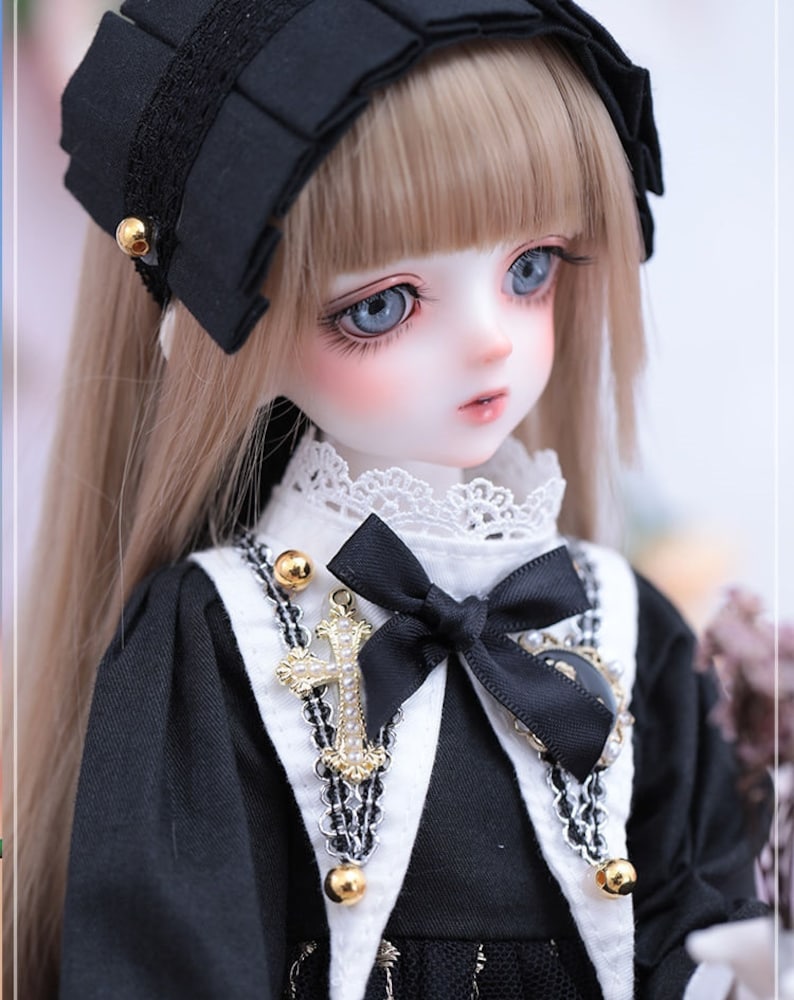 May include: A detailed portrait of a doll with long, light brown hair and blue eyes. The doll wears a black dress with a white collar and a black bow. A gold cross pendant and a black headband with gold accents complete the look.