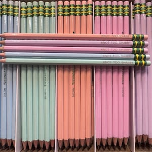 Puede incluir: Una colección de lápices de colores pastel, incluyendo azul claro, verde menta, melocotón, rosa y lavanda, dispuestos en una caja. Algunos lápices están etiquetados como "KNOT TOO SHABBY" y "DIXON TICONDEROGA".