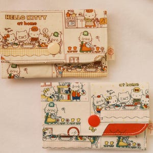 Może przedstawiać: Dwa portfele Hello Kitty w domu. Portfele są beżowe z powtarzającym się wzorem postaci Hello Kitty w otoczeniu kuchennym. Jeden portfel ma beżowe zapięcie na guziki, a drugi czerwone zapięcie na guziki.