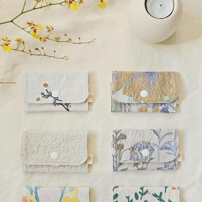 May include: Several small fabric wallets with diverse floral patterns and textures. Each wallet has a flap closure with a white button. Some are embroidered, others printed. The wallets are displayed on a cream-coloured surface, with a candle holder and flowers.