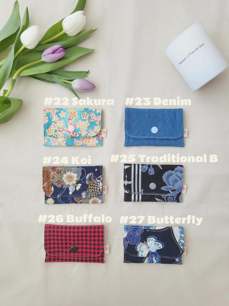 May include: A selection of small fabric wallets with diverse patterns and colours. Designs feature floral, denim, koi fish, buffalo plaid, and butterfly patterns. Each wallet has a button closure. The text "#22 Sakura", "#23 Denim", "#24 Koi", "#25 Traditional B", "#26 Buffalo", and "#27 Butterfly" is visible.