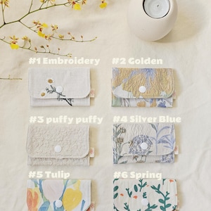 May include: Six small fabric wallets with various floral and textured patterns. Each wallet features a button closure. The wallets are labelled with the following text: #1 Embroidery, #2 Golden, #3 puffy puffy, #4 Silver Blue, #5 Tulip, and #6 Spring.