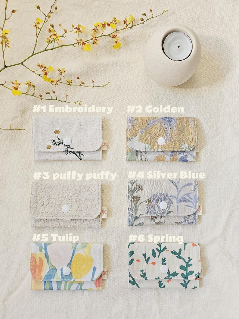 May include: Six small fabric wallets with various floral and textured patterns. Each wallet features a button closure. The wallets are labelled with the following text: #1 Embroidery, #2 Golden, #3 puffy puffy, #4 Silver Blue, #5 Tulip, and #6 Spring.