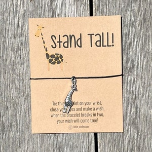 Peut inclure: Une carte marron avec une girafe de dessin animé et le texte "Stand Tall!" Un bracelet en cordon noir avec un charme de girafe argenté est attaché à la carte. La carte indique "Attachez ce bracelet à votre poignet, fermez les yeux et faites un vœu, lorsque le bracelet se cassera en deux, votre vœu se réalisera !"  little wishes.co