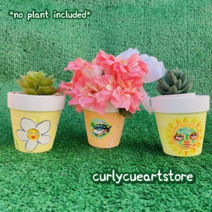 Pode incluir: Três vasos de flores pintados à mão com desenhos extravagantes. Os vasos são amarelos com bordas brancas e apresentam uma margarida, um olho e um rosto de sol. O texto "curlycueartstore" está na parte inferior e "*no plant included*" na parte superior.