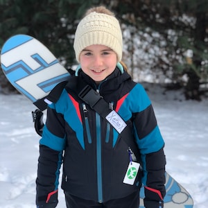 May include: A young person wearing a white knit hat and a blue and black ski jacket with a red stripe. They are holding a blue and white snowboard with a black strap.