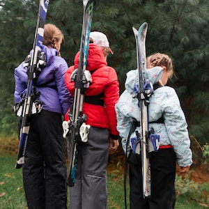 以下が含まれることがあります： 冬用のジャケットを着た3人の子供が、背中にスキーを背負っています。スキーは黒、青、白です。子供たちは緑色の松の木の前に立っています。