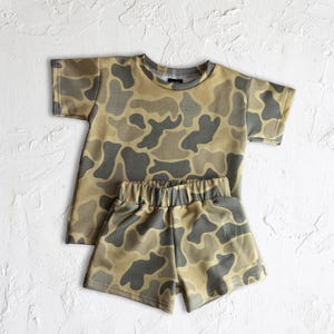May include: A two-piece children's outfit featuring a camouflage pattern in shades of tan, gray, and olive green. The set includes a short-sleeved t-shirt and matching elastic-waist shorts. The outfit is displayed on a textured white surface.