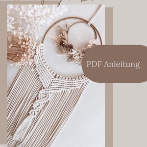 Op de afbeelding: Een beige macramé droomvanger met een houten ring en een PDF-instructiehandleiding. De droomvanger hangt aan een witte muur en heeft een kwast aan de onderkant.