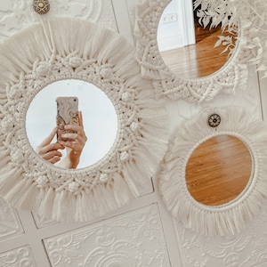 May include: Three white macrame mirrors with fringe hanging on a white wall with decorative molding. The mirrors are round and have a natural wood finish.