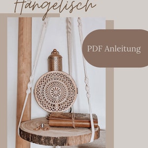 Peut inclure: Une étagère suspendue en macramé avec une tablette en bois circulaire retenue par des cordes nouées couleur crème. Elle supporte une lanterne métallique ronde aux motifs découpés, un petit livre en cuir et une bague. Texte : Makramee Hängetisch PDF Anleitung.