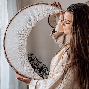 Puede incluir: Un colgante de pared con forma de media luna de macramé blanco, con un diseño geométrico y flecos. El marco es de metal dorado. Esta pieza decorativa es de estilo bohemio, ideal para la decoración de paredes.