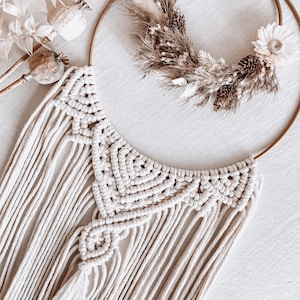 May include: A white macrame wall hanging with a geometric design and a tassel. The hanging is attached to a gold metal hoop with dried flowers and grasses.