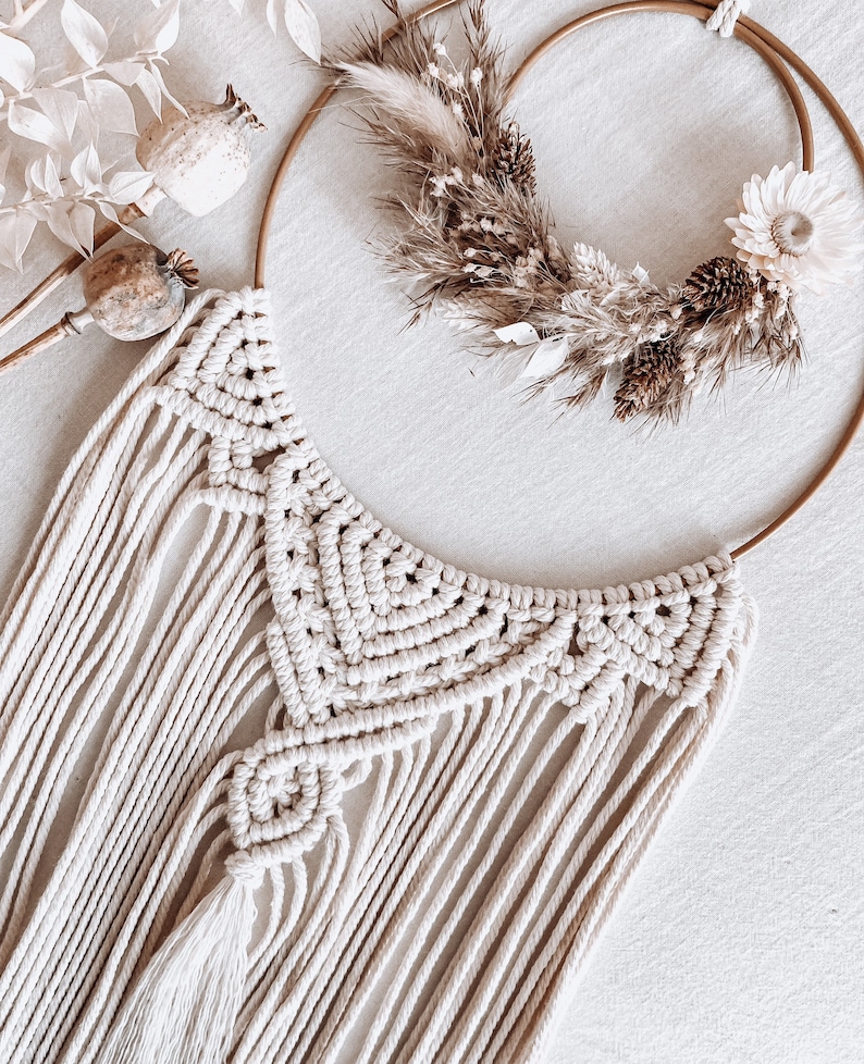 May include: A white macrame wall hanging with a geometric design and a tassel. The hanging is attached to a gold metal hoop with dried flowers and grasses.
