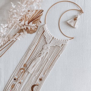 Può includere: Un arazzo da parete in macramè bianco a forma di mezzaluna, con dettagli dorati e un fiocco. L'arazzo è decorato con piccole stelle e lune dorate.