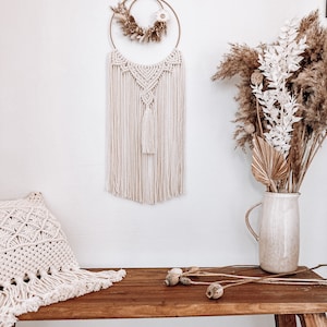 May include: A macrame wall hanging with a wooden ring and dried flowers. The wall hanging is cream coloured and has a tassel at the bottom. The wall hanging is hanging on a white wall. A wooden bench with a cream coloured pillow is in the foreground.