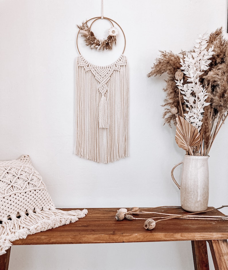 May include: A macrame wall hanging with a wooden ring and dried flowers. The wall hanging is cream coloured and has a tassel at the bottom. The wall hanging is hanging on a white wall. A wooden bench with a cream coloured pillow is in the foreground.