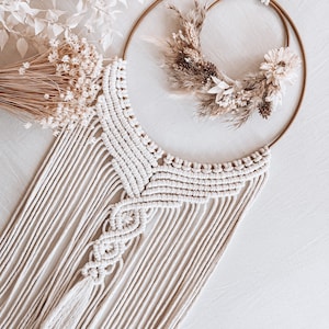 Könnte beinhalten: Ein Makramee-Wandbehang mit einem cremefarbenen Quasten und einem Doppelring-Kranz mit getrockneten Blumen. Der Kranz ist aus natürlichen Materialien gefertigt und hat einen rustikalen, bohemian Stil.