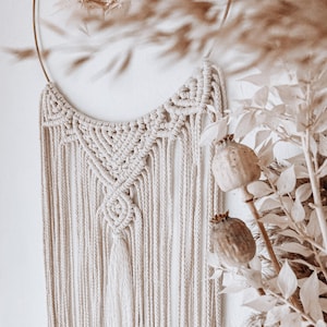 May include: A white macrame wall hanging with a geometric design and fringe. The hanging is made of cotton cord and is attached to a gold metal hoop.
