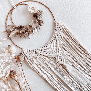May include: A white macrame wall hanging with a gold hoop and dried floral accents.