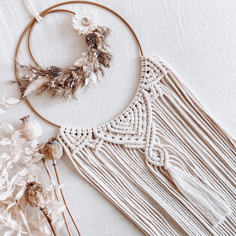 May include: A white macrame wall hanging with a gold hoop and dried floral accents.