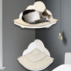 Op de afbeelding: Twee witte, wolk-vormige kattenplanken aan een grijze muur. De bovenste plank heeft een slapende kat met zwart-witte markeringen. De planken hebben beige kussens. Links is een raam met een plant zichtbaar.