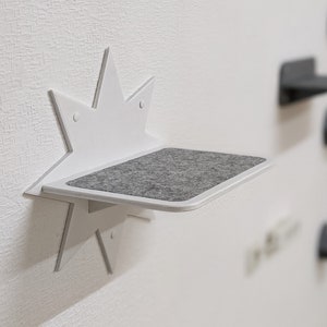 May include: White star-shaped cat shelf with a gray felt surface. The shelf is mounted on a white wall.