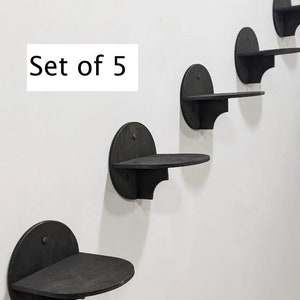 May include: Set of five black wooden wall shelves in a staggered formation. The shelves are rounded and have a slight lip on the front edge.  "Set of 5" is written in white text on a white background.