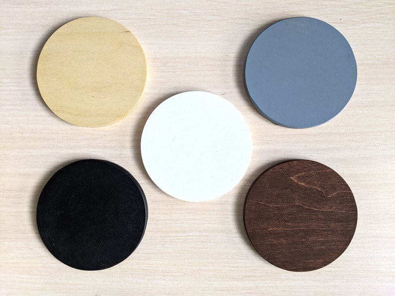 May include: Five round wooden coasters in different colors: natural wood, white, black, gray, and dark brown.