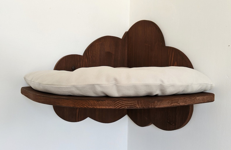May include: A wooden cat bed shaped like a cloud with a beige cushion. The bed is mounted on a wall and is perfect for a cat to sleep or relax on.