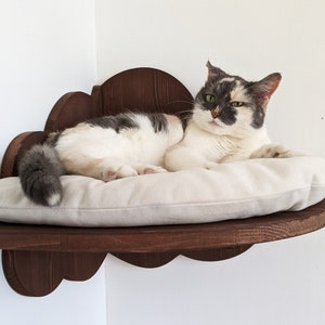 May include: A white and gray cat with green eyes is lying on a white cushion in a wooden cloud-shaped cat bed mounted on a wall.