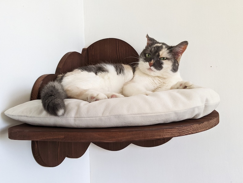 May include: A white and gray cat with green eyes is lying on a white cushion in a wooden cloud-shaped cat bed mounted on a wall.