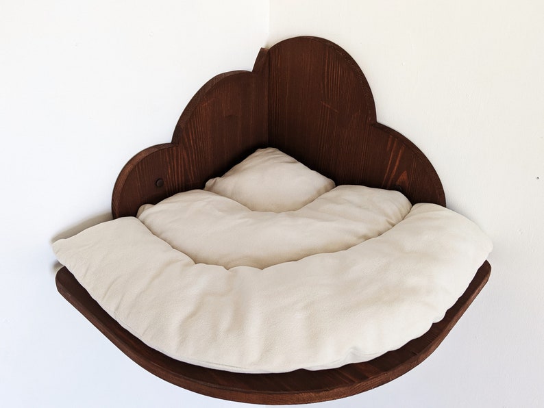 May include: A wooden cat bed shaped like a cloud, with a white cushion inside. The bed is mounted on a wall corner.