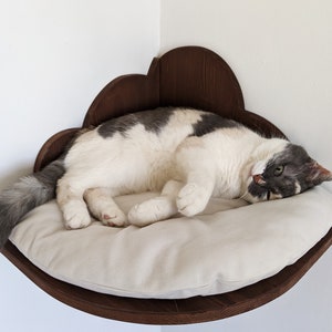 May include: A white and gray cat is sleeping in a brown wooden cat bed shaped like a cloud. The bed is mounted on a wall and has a soft, beige cushion.