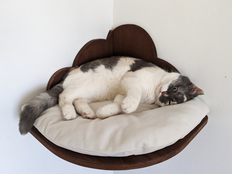 May include: A white and gray cat is sleeping in a brown wooden cat bed shaped like a cloud. The bed is mounted on a wall and has a soft, beige cushion.