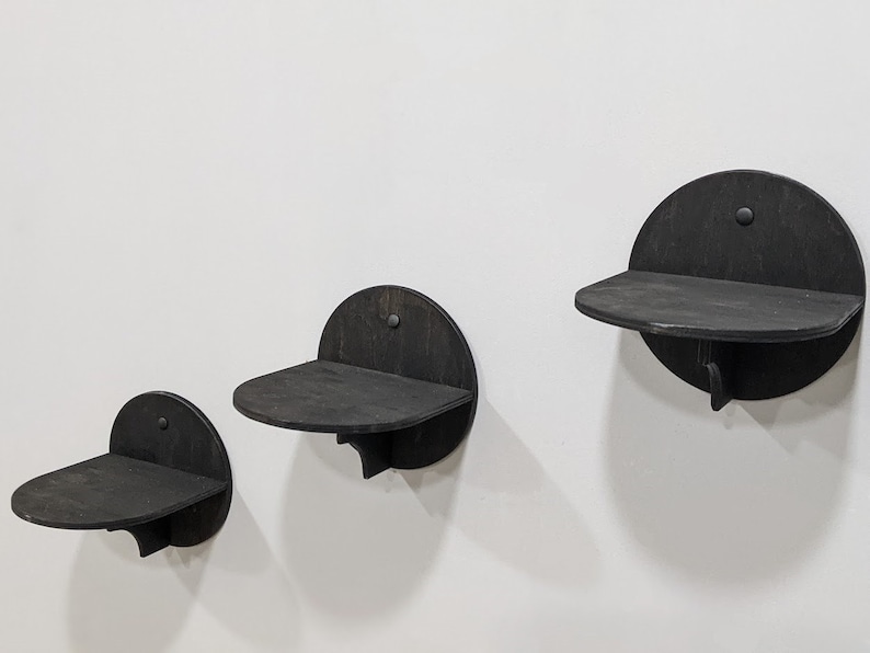 May include: Three black wooden wall shelves in different sizes. The shelves are semi-circular and have a slight lip on the front edge. The shelves are mounted on a white wall.