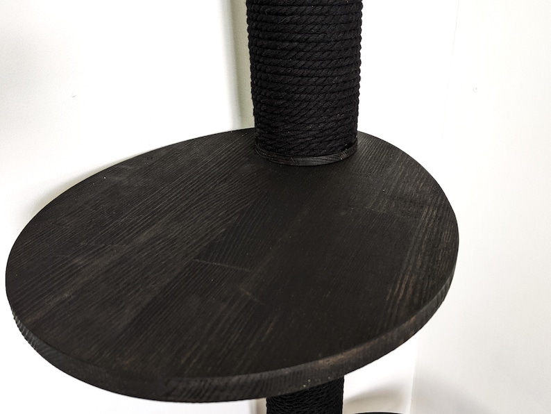May include: A black wooden platform with a black rope-covered post attached to it. The platform is round and has a slightly raised edge.
