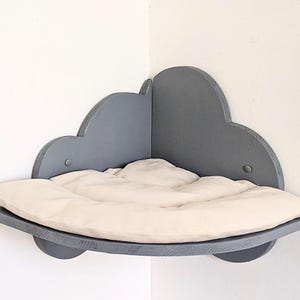 Op de afbeelding: Een grijs hoekkattenbed in de vorm van een wolk met een beige kussen. Het bed is aan een muur bevestigd.