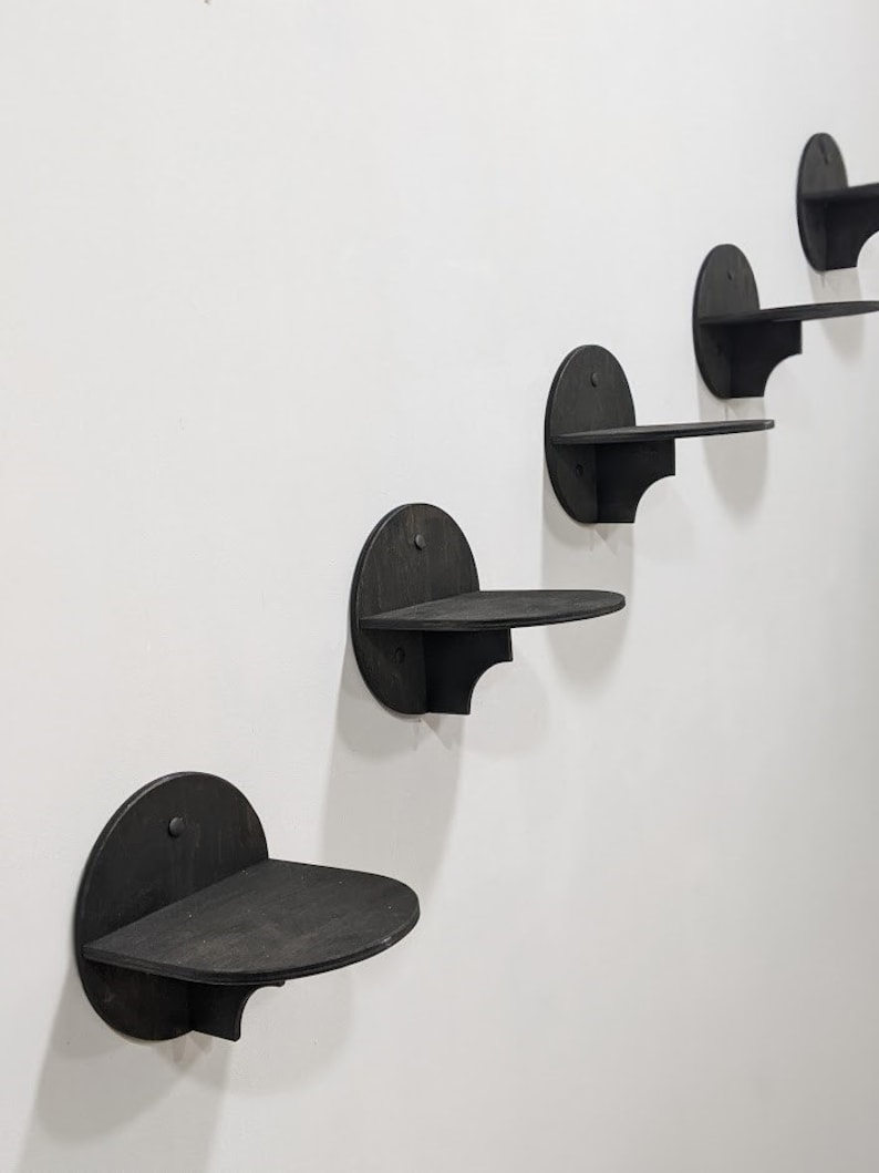 May include: Five black wooden wall shelves in a staggered formation. Each shelf is a semi-circle with a flat surface and a curved edge.