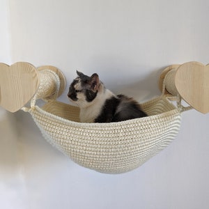 May include: A white crocheted cat hammock with a black and white cat sitting inside. The hammock is attached to two wooden heart-shaped wall mounts with sisal scratching posts.