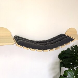 May include: A wooden cat hammock with a gray cushion, attached to two wooden shelves. The hammock is suspended between the shelves with rope.