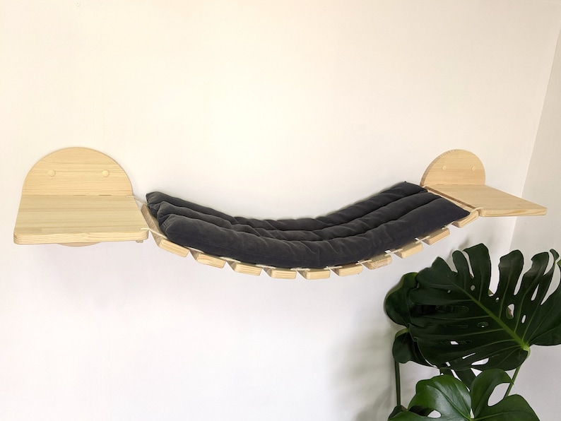 May include: A wooden cat hammock with a gray cushion, attached to two wooden shelves. The hammock is suspended between the shelves with rope.