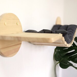 May include: A wooden cat bed shelf with a black cushion, mounted on a white wall. The shelf is curved and has a circular base.