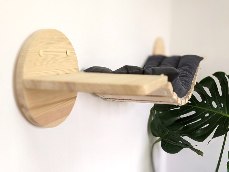 May include: A wooden cat bed shelf with a black cushion, mounted on a white wall. The shelf is curved and has a circular base.