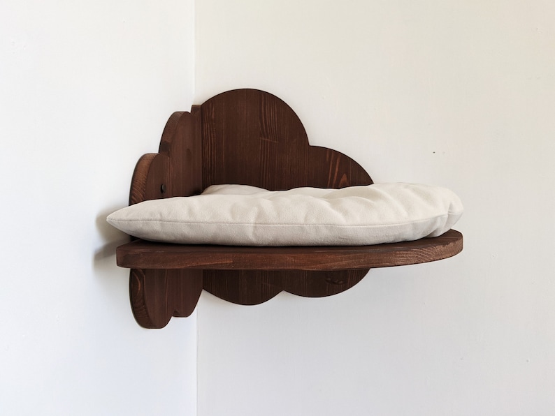 May include: A wooden cat bed shaped like a cloud, with a beige cushion. The bed is mounted on a corner wall.