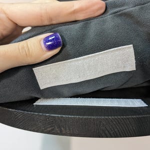 May include: Close-up of a black fabric cushion with two white hook and loop fasteners. The cushion is being held by a hand with purple nail polish.